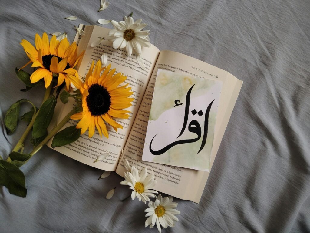 Flat lay of an open book with sunflowers and Arabic script card on a grey background.